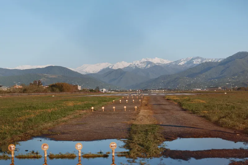 A rural runway stretches into the distance with small runway lights on both sides and snow-capped mountains visible under a clear blue sky.