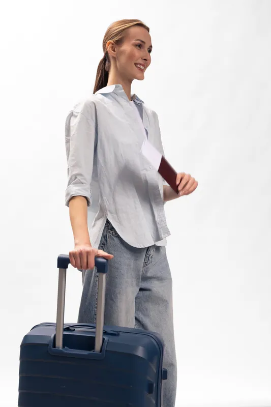 Smiling woman in casual clothes holds passport and wheeled suitcase standing on white background. Concept for travel, tourism, vacation, and business trip preparation.