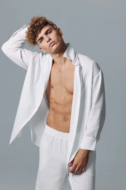 Young man with curly hair wearing white open shirt and pants posing with hand on head against gray background. Fashion model with casual style and confident expression in studio.
