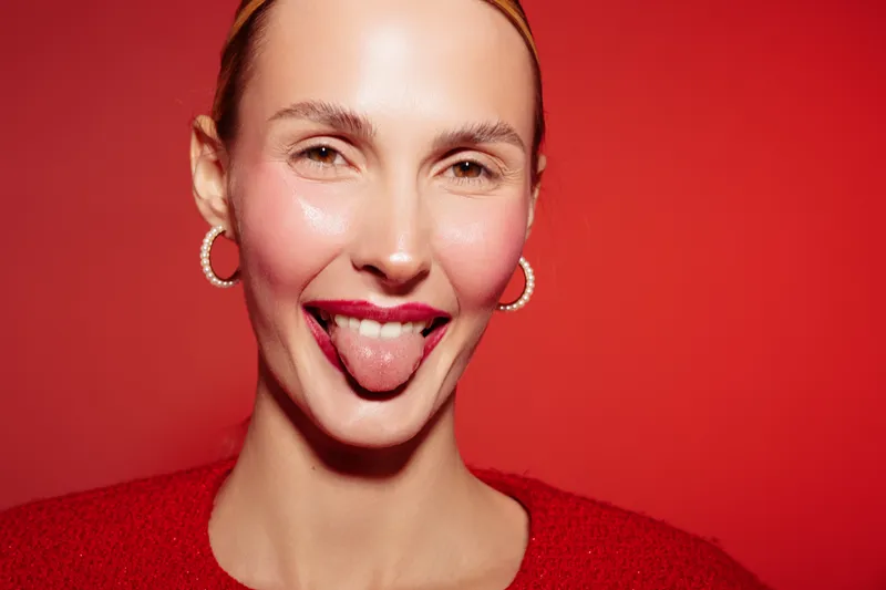 Young woman with clear skin and red lipstick sticks out tongue playfully against red background. Concept of fun, beauty, expression, and youthful confidence in studio portrait.