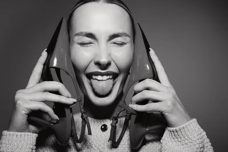 Happy Caucasian woman with light skin holding high heel shoes near her face, smiling with eyes closed and tongue out in black and white studio photo.