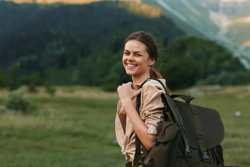 Young woman traveler with backpack smiles happily standing in green meadow with mountains in background enjoying outdoor lifestyle and nature adventure.