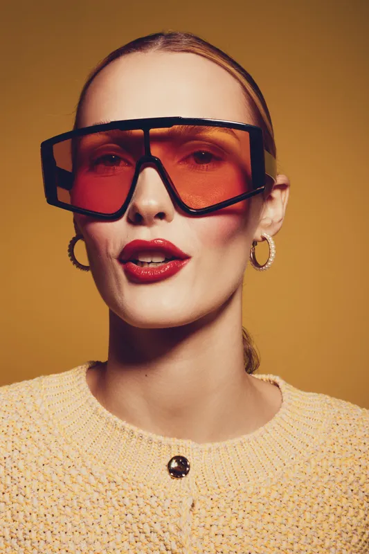 Stylish woman with red lipstick wearing oversized orange sunglasses and hoop earrings poses against warm yellow background. Fashion portrait with modern accessories and confident expression.