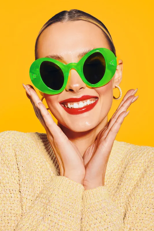 Smiling woman with bright red lipstick wearing large green sunglasses and a beige sweater on yellow background. Concept of fashion, summer style, and joyful mood.