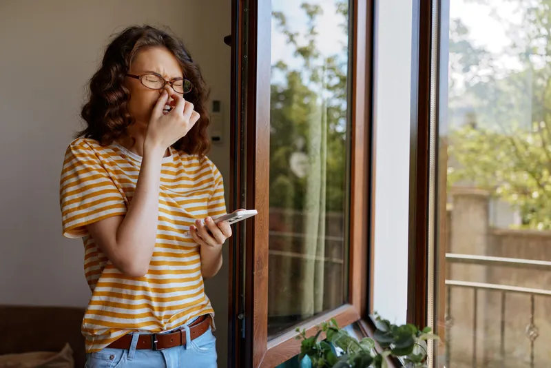 Smiling woman with curly hair wearing a striped shirt stands by a window, wiping her nose with tissue while looking at a phone. Bright natural light fills the cozy home, capturing a casual morning