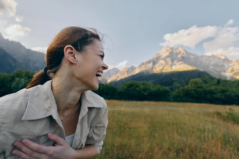 Happy woman outdoors laughing with eyes closed, natural light, casual clothing, mountain landscape background, green grass field, relaxed lifestyle moment