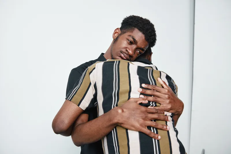 Two young black men hugging each other in emotional support with dark skin and casual clothing on white background for friendship and comfort.