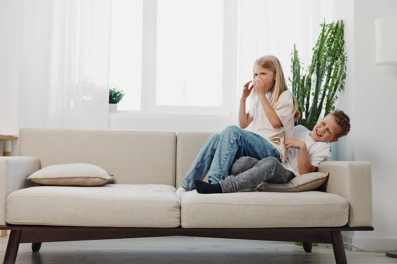 Happy siblings laughing and playing on a beige sofa in a bright living room with natural light and green plants. Concept of family fun, childhood joy, and home comfort.