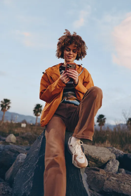 Woman lifestyle outdoors selfie, curly hair, warm jacket and denim, holding phone on rocky trail at sunset, calm confident smile, soft breeze, natural landscape background
