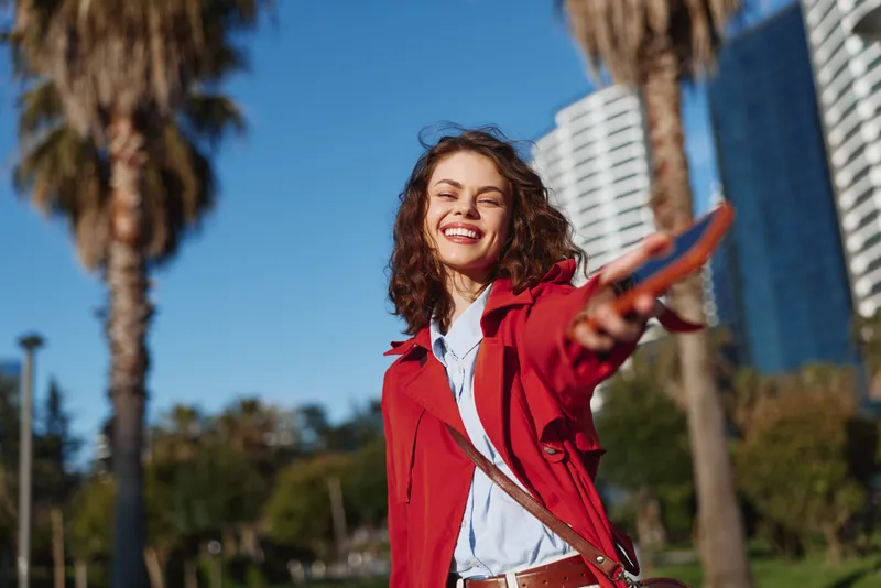 Bright young woman outdoors wearing a red jacket smiles at the camera in a sunny city park with palm trees, tall buildings, and a clear blue sky behind, a gentle breeze.