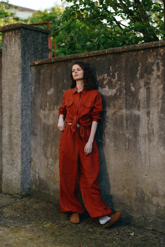 A stylish woman in a red jumpsuit stands beside a weathered wall in an outdoor setting. A calm, confident lifestyle moment captured in daylight