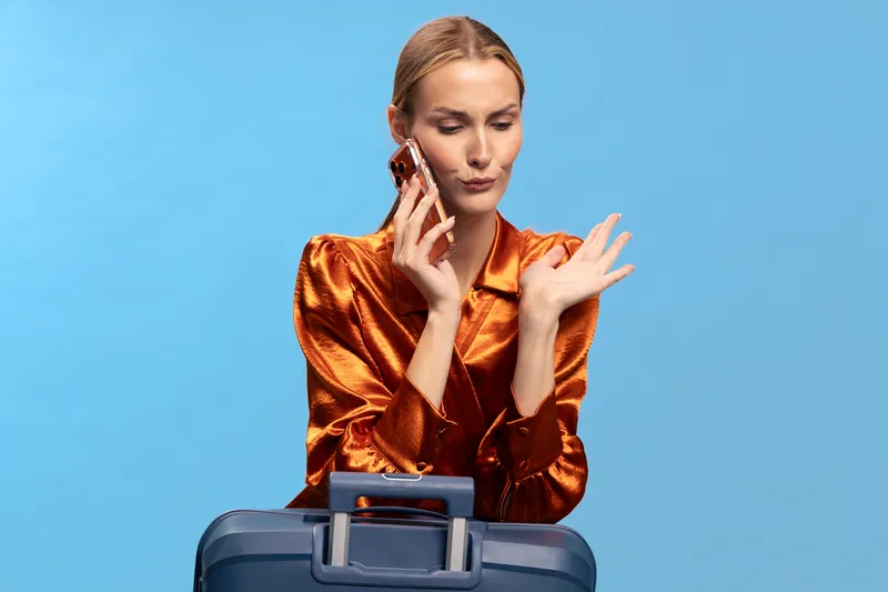 Young woman in orange satin blouse talks on smartphone with skeptical expression, sitting behind blue suitcase on blue background. Concept for travel, communication, doubt, and business trip.