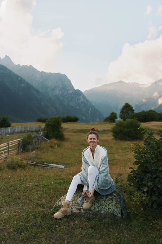Casual joyful woman sits on a rock in a scenic mountain meadow, wearing a cardigan and white jeans, smiling, enjoying outdoor life in a peaceful rural landscape under soft daylight.