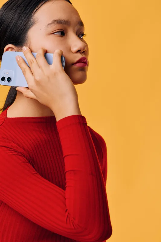 Young woman wearing red sweater holding phone to ear for conversation with serious expression on orange background in studio. Communication, connection, technology concept for mobile call.