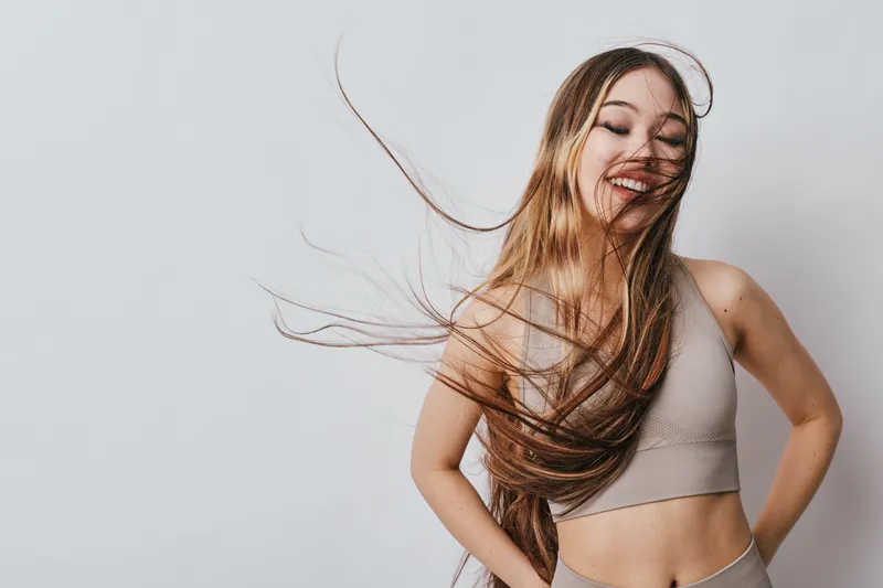 Young woman with long hair wearing sportswear stands on light background smiling with closed eyes in relaxed pose. Fitness, health and happy lifestyle concept.