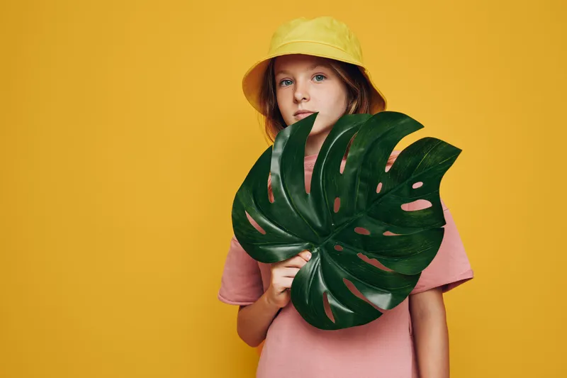 Young girl wearing a yellow hat and pink shirt holding a large green tropical leaf against a yellow background. Studio portrait with bright vibrant colors.