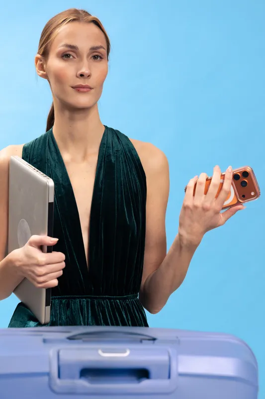 Young woman in elegant dress holds laptop and smartphone near suitcase on blue background. Concept for travel, business, technology, and modern lifestyle with confident expression.