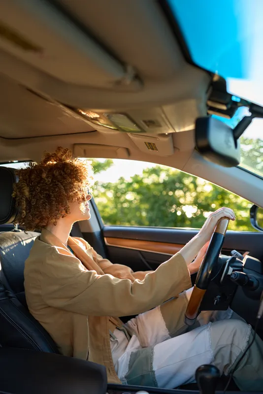 Cheerful woman driving car, sunlit interior, relaxed posture, casual outfit, warm tones, natural light, soft breeze, green landscape visible through window