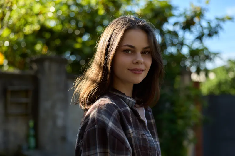 Casual woman lifestyle portrait outdoors, smiling softly in plaid shirt, natural sunlight, green garden background, relaxed pose capturing everyday confidence and calm mood