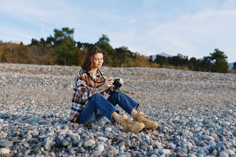 Woman photographer sits on a pebble beach, holds a camera and photographs the scenic shoreline during daylight, casual plaid shirt and boots, calm natural setting.