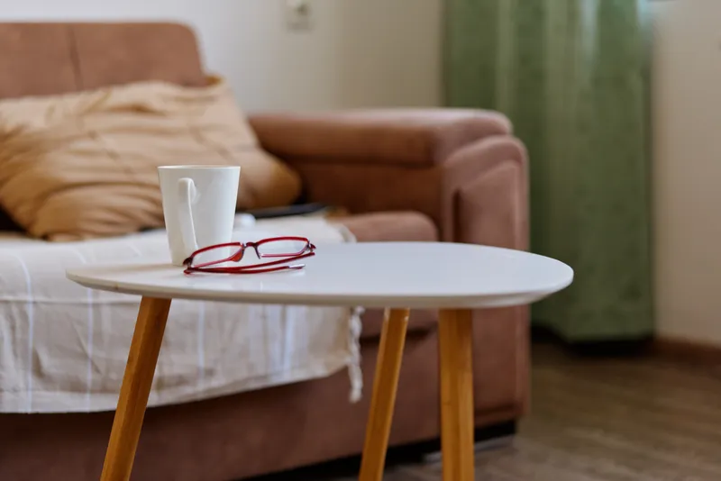 Cozy living room scene with a round white table, a mug and glass resting nearby, a soft sofa in the background, a warm natural light creating a calm home atmosphere soft textures in view.