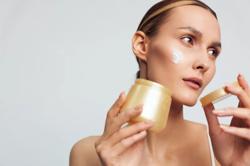 Young woman with sleek hair applies cream from a gold jar on her cheek in a bright studio, showcasing skincare routine and self care with soft lighting that creates a calm, confident mood.