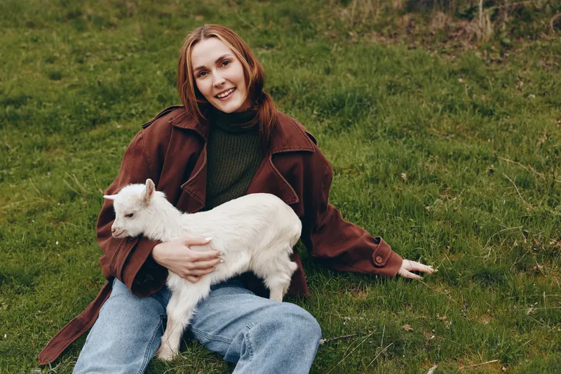Woman outdoors with a goat kid, smiling, sits on grass, wearing a brown coat and green sweater, a warm lifestyle moment of affection and companionship with animals