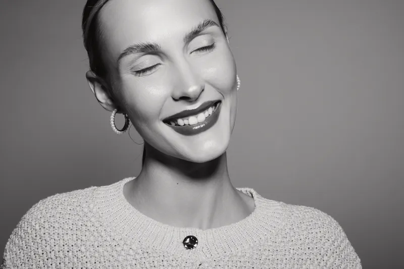 Happy Caucasian woman with light skin closes eyes and smiles softly. She wears earrings and a knitted sweater against plain background. Concept of joy, beauty, and positivity.