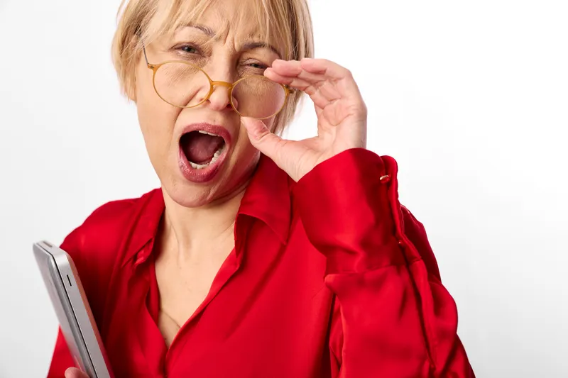 Woman, elderly, glasses, laughing, surprised, red blouse, studio white background, holding eyewear frame with tissue near mouth, expressive face, closeup indoor moment, cheerful emotion