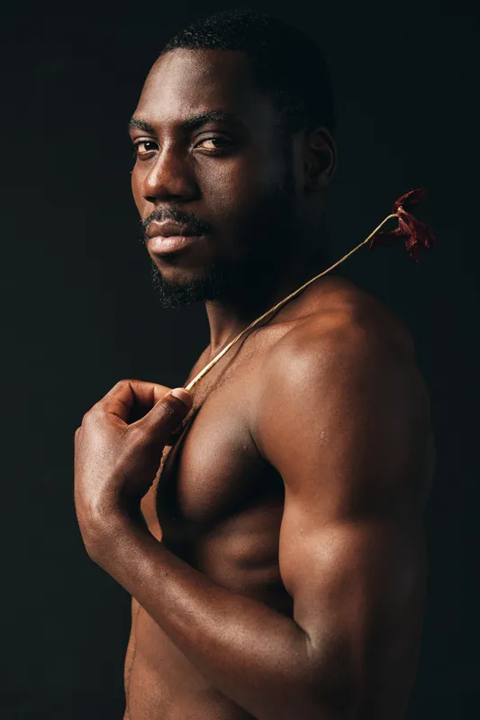 African man with muscular body and bare torso holding dried flower on shoulder in creative conceptual visual style with dark background and dramatic lighting.