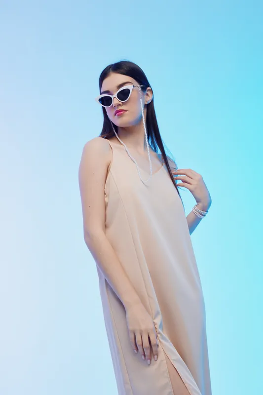 Young woman in a beige dress wearing white sunglasses poses confidently against a blue background. Concept for beauty, fashion, and modern style.