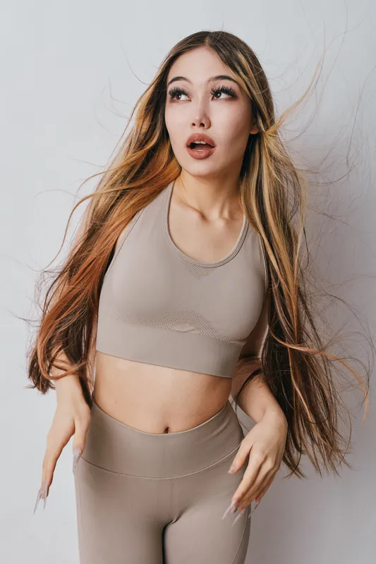 Young woman in beige activewear posing with long hair looking surprised or thoughtful against light background in studio interior.