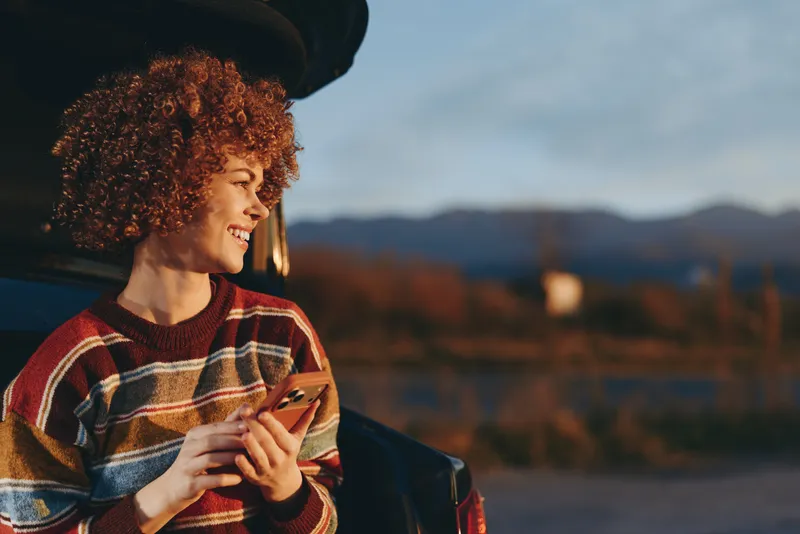 Lifestyle portrait of a woman outdoors at sunset, curly hair and smile, holding a small drink near her bag, travel mood with mountains landscape, open road, soft evening light
