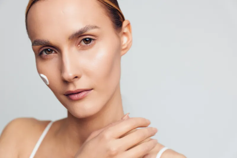 Young woman with clean skin applies moisturizing cream on her face, showing natural beauty and skincare routine in closeup portrait with calm expression and neutral background.