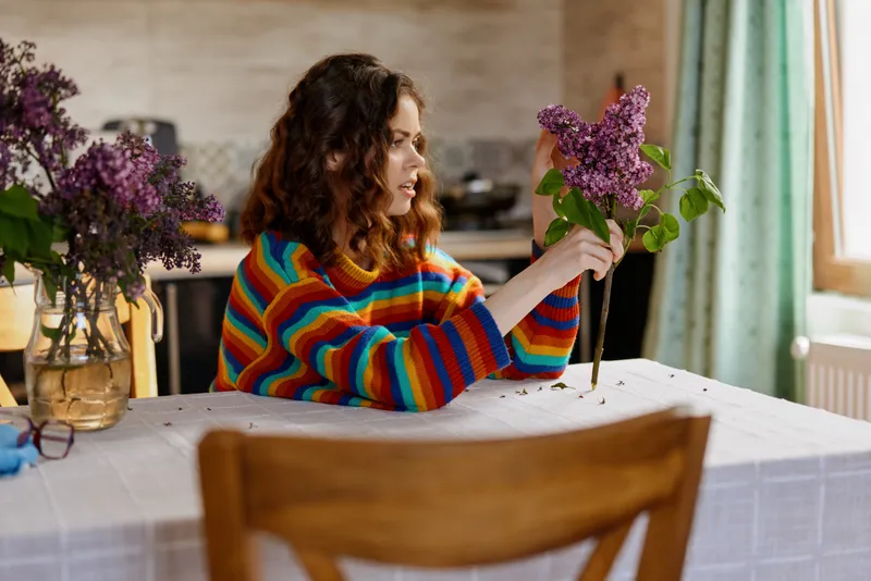 Candid woman indoors at a sunlit table, holding a bouquet of flowers, wearing a colorful striped sweater, capturing a relaxed lifestyle moment in a cozy home setting.