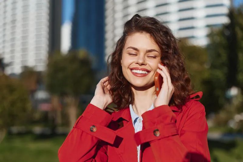 Cheerful woman outdoors, smiling while talking on a phone, red jacket adds color against a sunny urban park background, bright natural light, carefree atmosphere