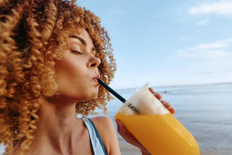 Portrait of a woman with curly hair sipping orange juice through a straw at the beach on a sunny day, showcasing a relaxed summer mood and coastal scenery in the background.