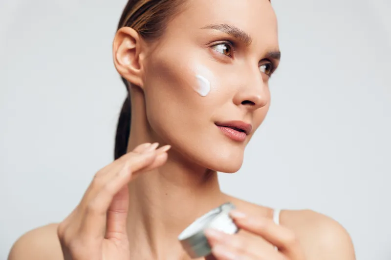Young woman applying white face cream on her cheek with soft natural makeup. Close-up portrait on light background. Concept of skincare, beauty routine, and healthy skin care.