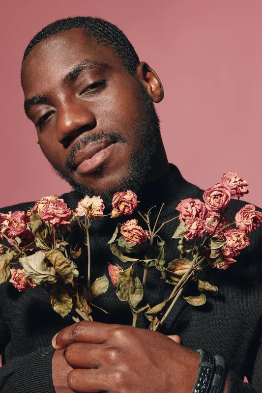 African man with dark skin holding dried pink roses in creative conceptual style against pink background. Portrait with closed eyes and peaceful expression in studio lighting.