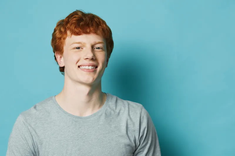 Young man with red hair and freckles smiling in gray t shirt on blue background, portrait of happy teenager with casual style and confident expression indoors.