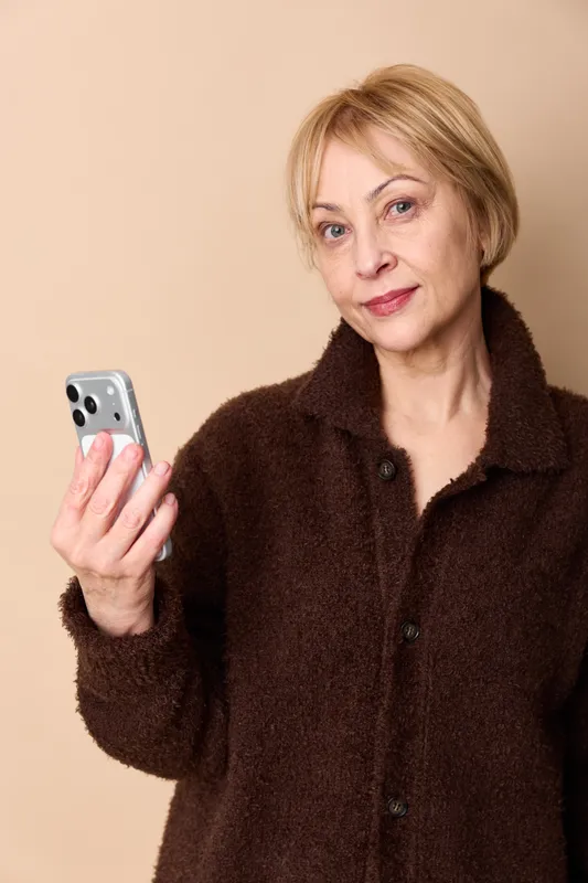 Woman smartphone portrait mature indoors communication texting call selfie technology moment, holding phone near face, brown coat, short blond hair, warm beige background