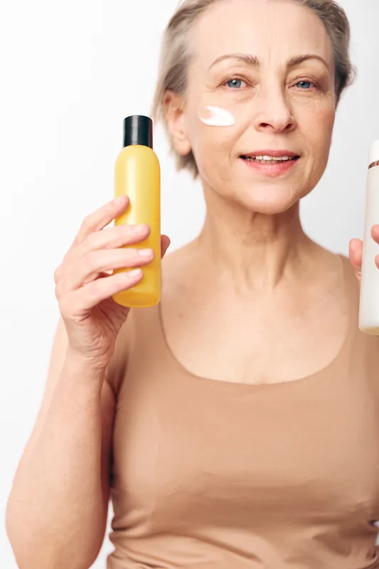 Mature woman with natural skin applies cream on face holding two cosmetic bottles. Healthy skincare routine concept for aging, beauty, wellness, and self-care in a bright studio setting.