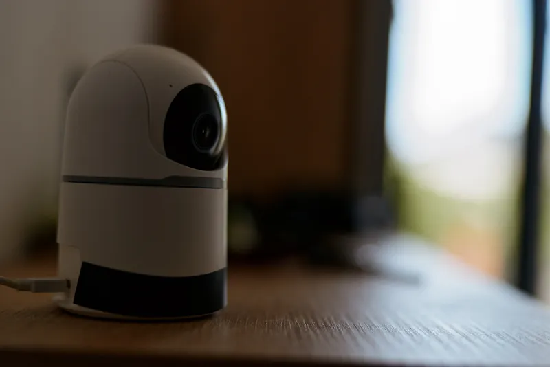 A compact indoor security camera sits on a wooden table, surveying a quiet living room with soft natural light and minimal decor.