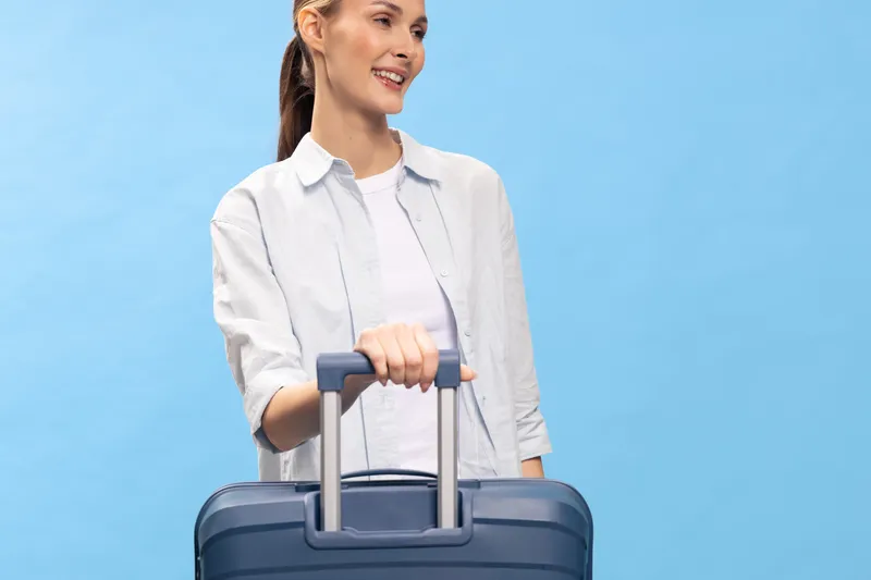 Smiling woman with ponytail stands holding a navy blue suitcase handle on a light blue background. Concept of travel, vacation, and modern lifestyle with casual white and grey clothing.