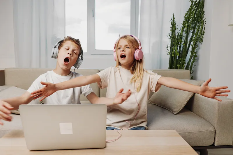 Two children wearing headphones sing passionately in front of a laptop on a wooden table in a bright living room with a sofa and plants. Concept of home entertainment and music fun.