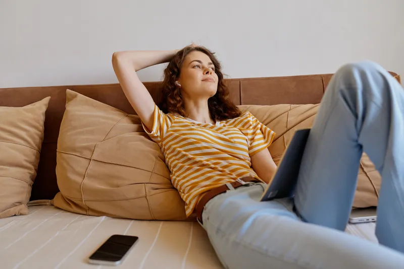A relaxed woman lounges on a couch at home with a laptop on her lap, a smartphone nearby. A casual lifestyle moment highlighting comfort, downtime, and home work balance.