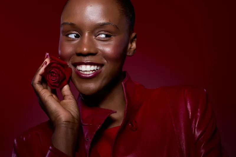 Smiling black woman with dark skin holds red rose near face, wearing red leather jacket. Studio portrait with burgundy background. Concept of beauty, fashion, and elegance.