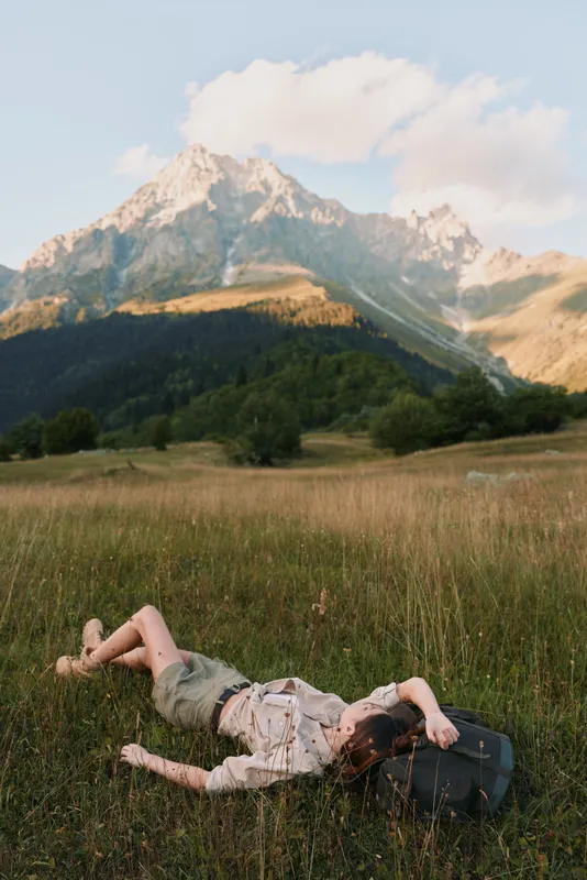 Woman in a lifestyle shot lies in a sunlit meadow with distant mountains, outdoor nature scene, relaxed mood and scenic open space