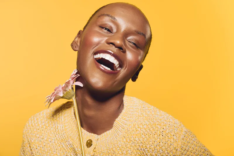 Happy smiling black woman with dark skin and short hair wearing yellow sweater holds pink flower against yellow background. Concept of joy, beauty, natural, and positive mood.