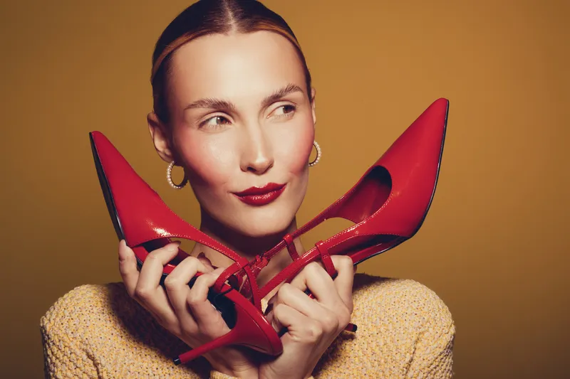 Young woman with slicked hair holds red high heel shoes near face, wearing yellow sweater and gold hoop earrings. Studio portrait on warm brown background with fashion and style concept.